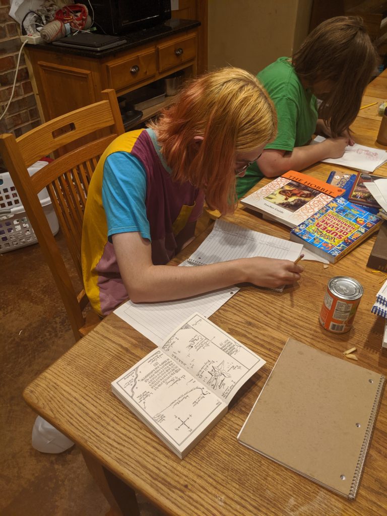 Blond teenager copying a map inscribed with runes from The Hobbit, another teen writing, table covered with books and papers